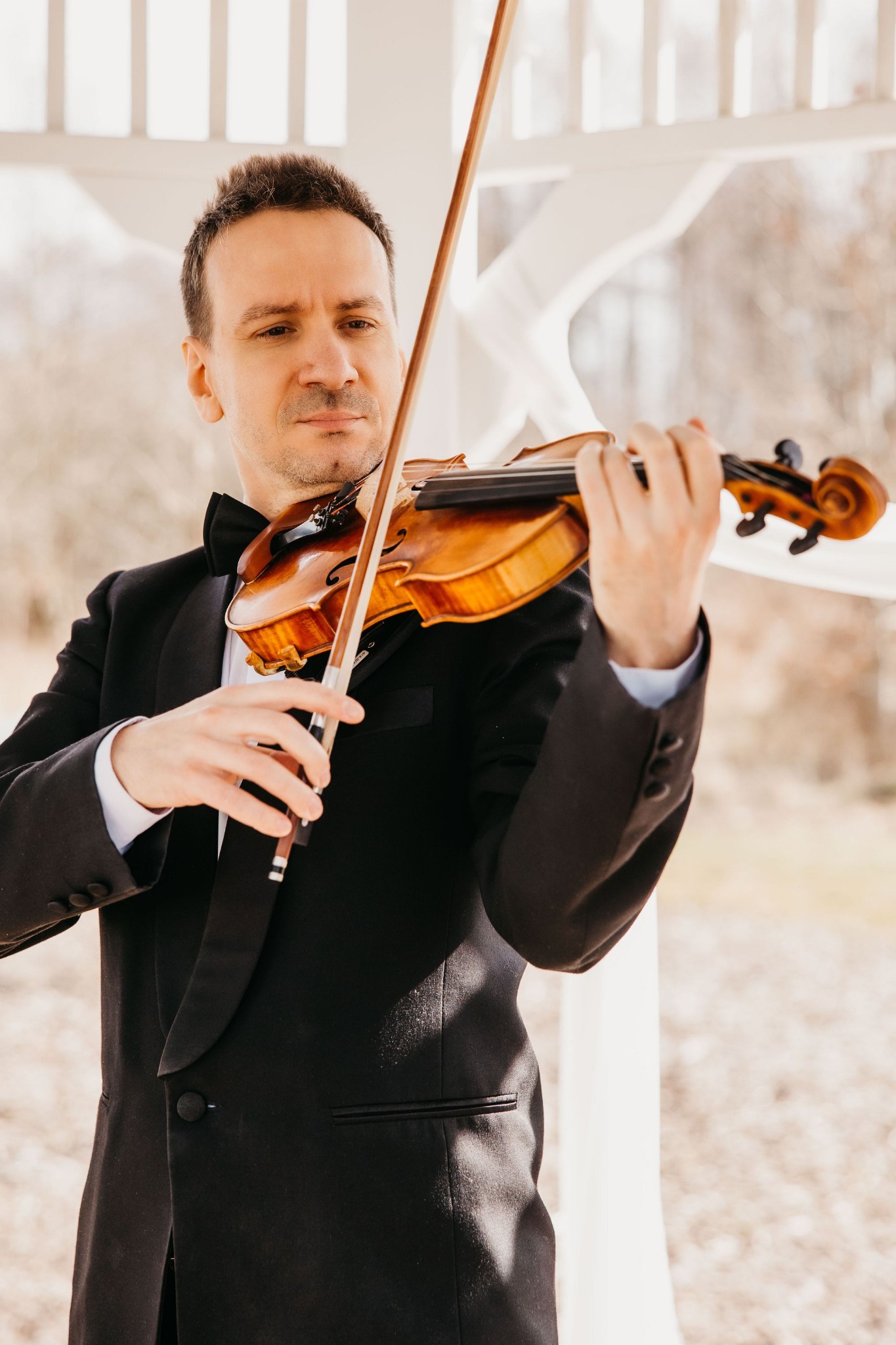 Mężczyzna w smokingu grający na skrzypcach w białej altanie.