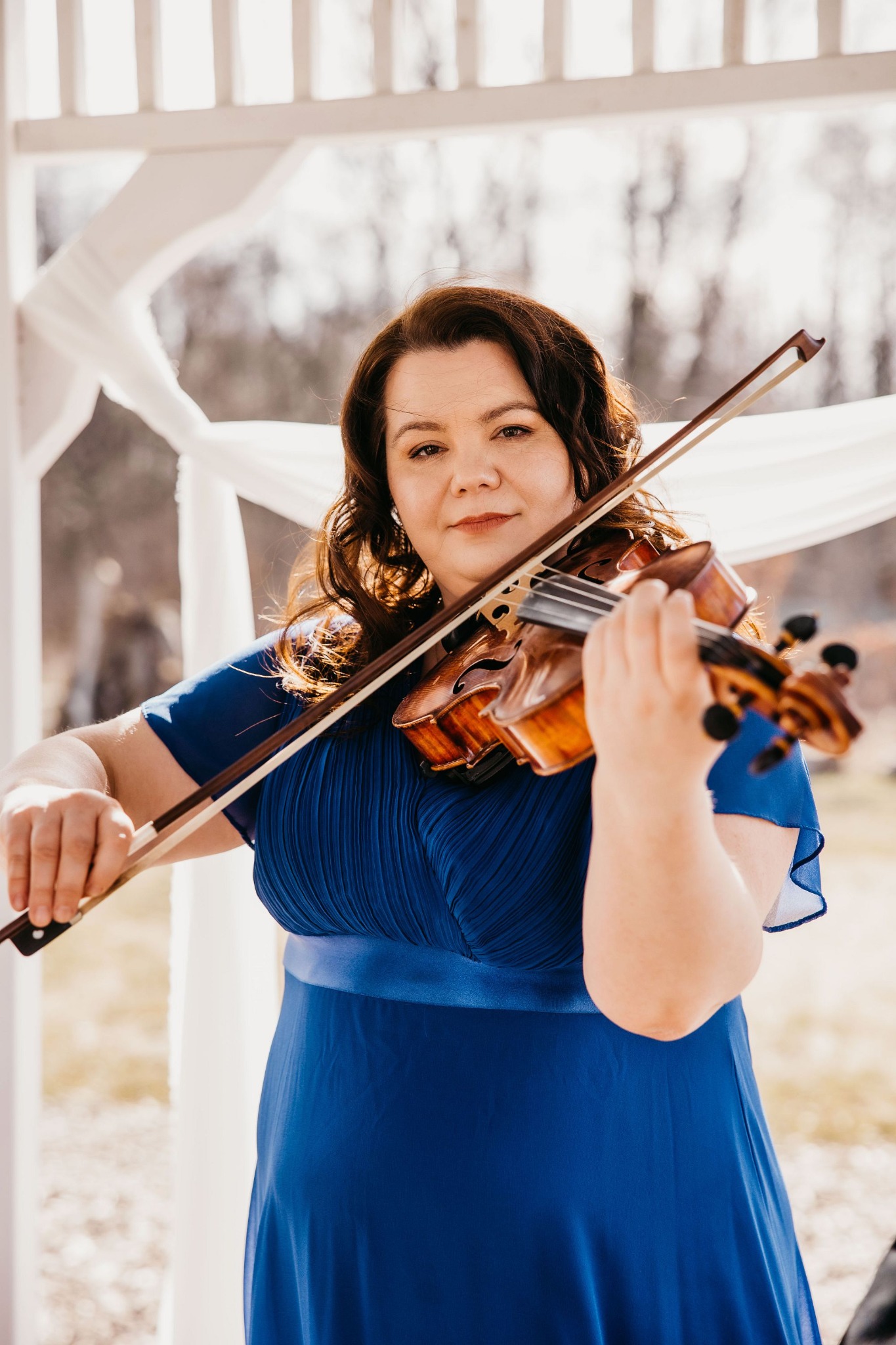 Kobieta w niebieskiej sukni gra na skrzypcach w białej altanie z widokiem na drzewa.