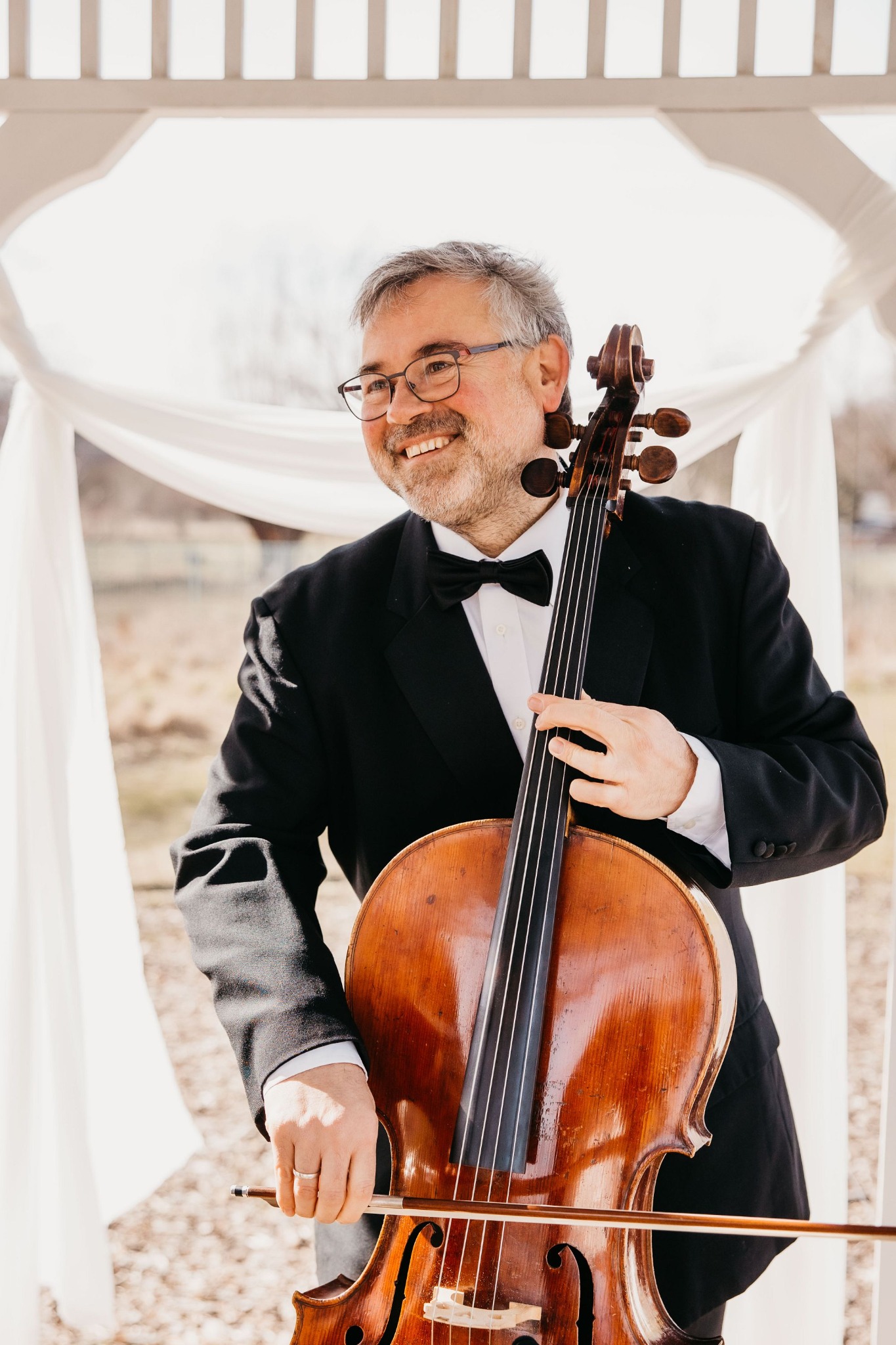 Uśmiechnięty mężczyzna w smokingu i muszce trzyma wiolonczelę przed białą altaną z draperiami.