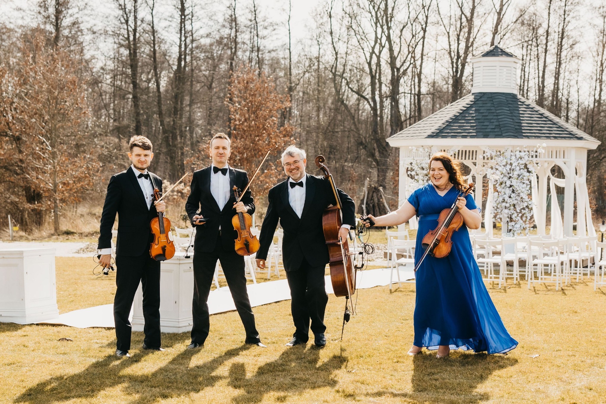 Kwartet smyczkowy w smokingach i sukni, z instrumentami, pozuje na trawniku przed białą altaną udekorowaną kwiatami, przygotowaną do ceremonii ślubnej.