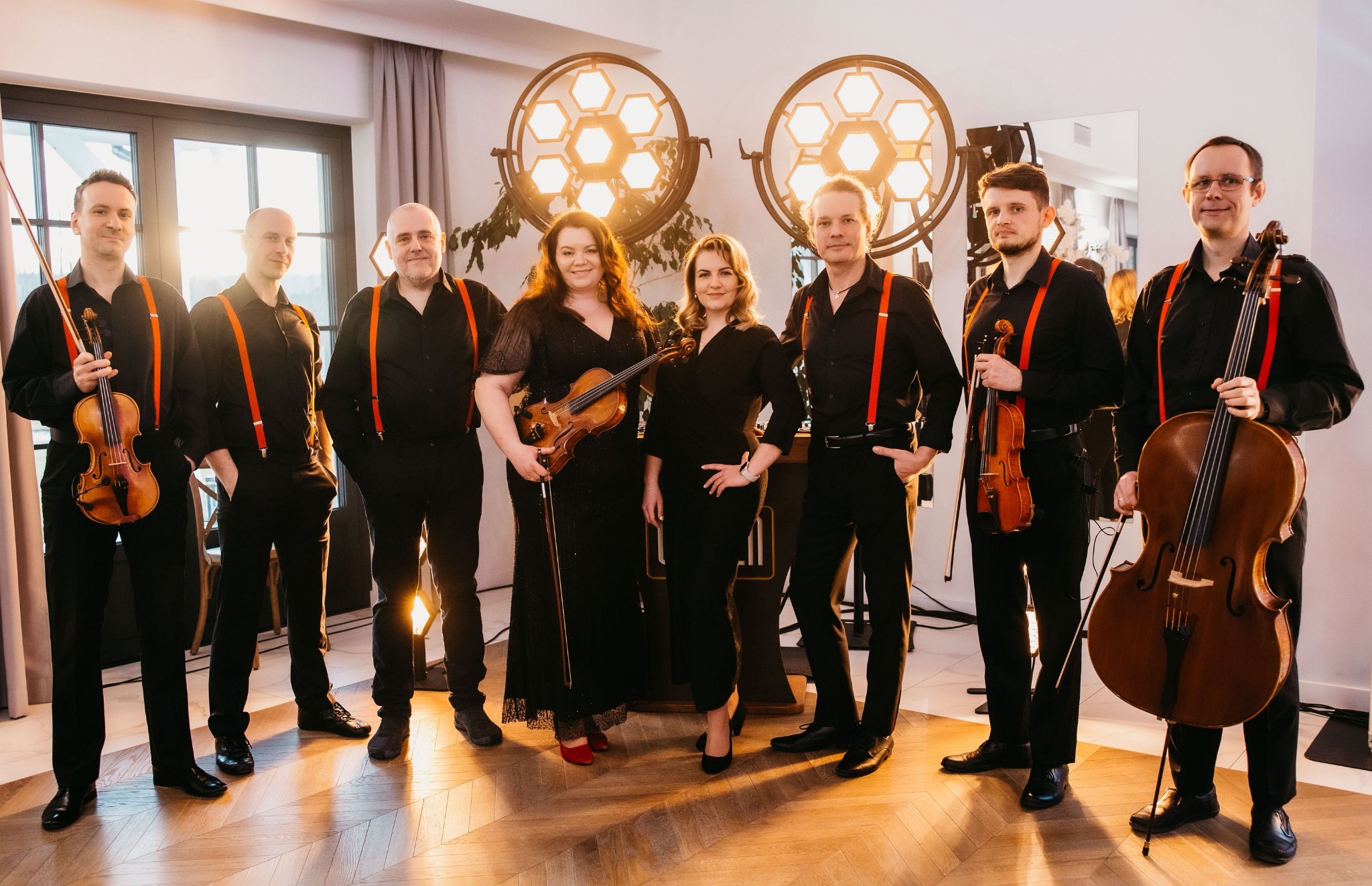 Ośmioosobowy zespół smyczkowy w czarnych strojach z czerwonymi szelkami, stojący na parkiecie w oświetlonym wnętrzu z dekoracyjnymi lampami w tle.