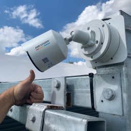 SKT PRO Kacper Till - Zewnętrzna kamera monitoringu Hikvision zamontowana na metalowej konstrukcji, widok z dołu na tle błękitnego nieba z chmurami, kciuk do góry.