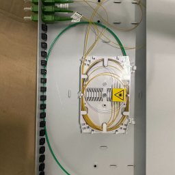 SKT PRO Kacper Till - Otwarta skrzynka światłowodowa z zielonymi złączami, żółtymi kablami światłowodowymi i naklejką ostrzegawczą o promieniowaniu laserowym wewnątrz.