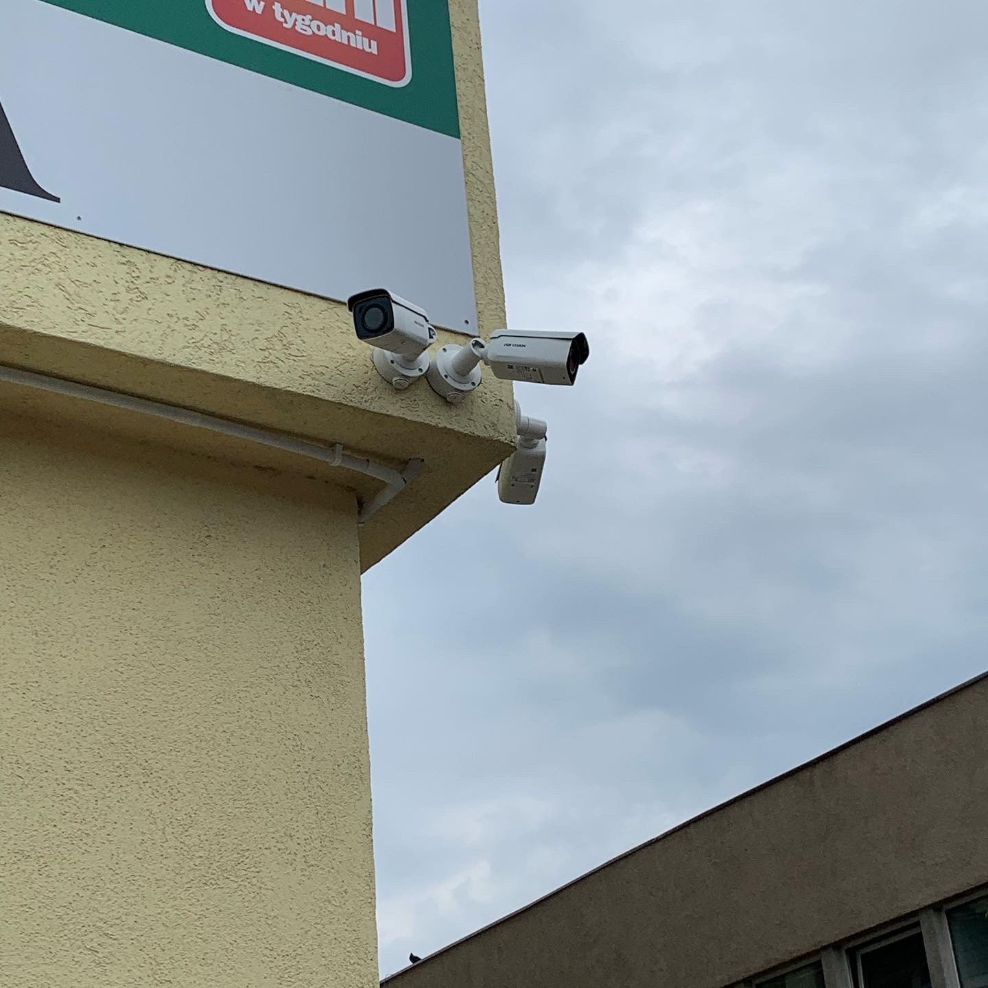 Dwie kamery monitoringu, jedna z obiektywem skierowanym w lewo, druga w prawo, zamontowane na rogu budynku z żółtą, teksturowaną elewacją pod pochmurnym niebem.