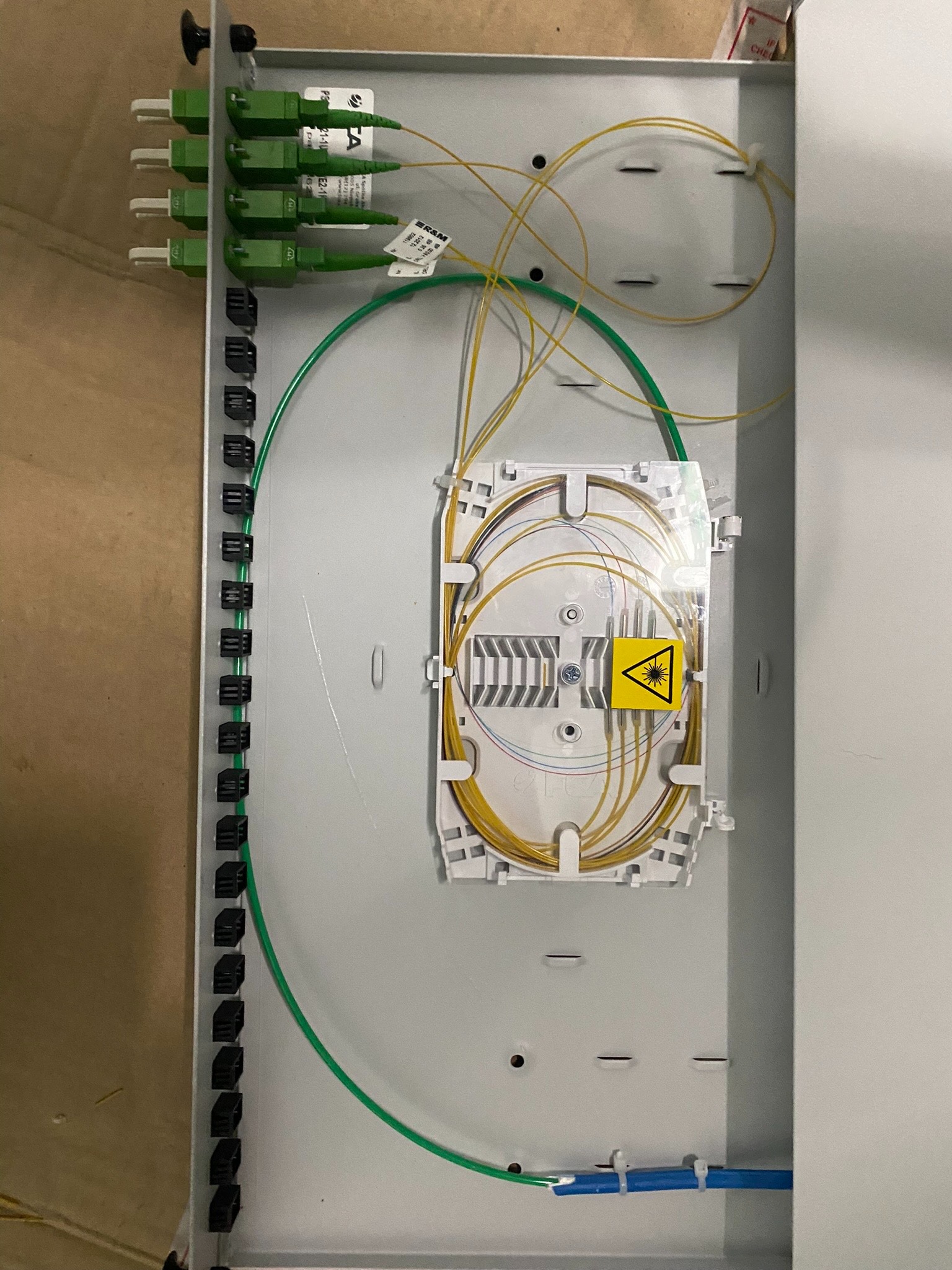 Otwarta skrzynka światłowodowa z zielonymi złączami, żółtymi kablami światłowodowymi i naklejką ostrzegawczą o promieniowaniu laserowym wewnątrz.