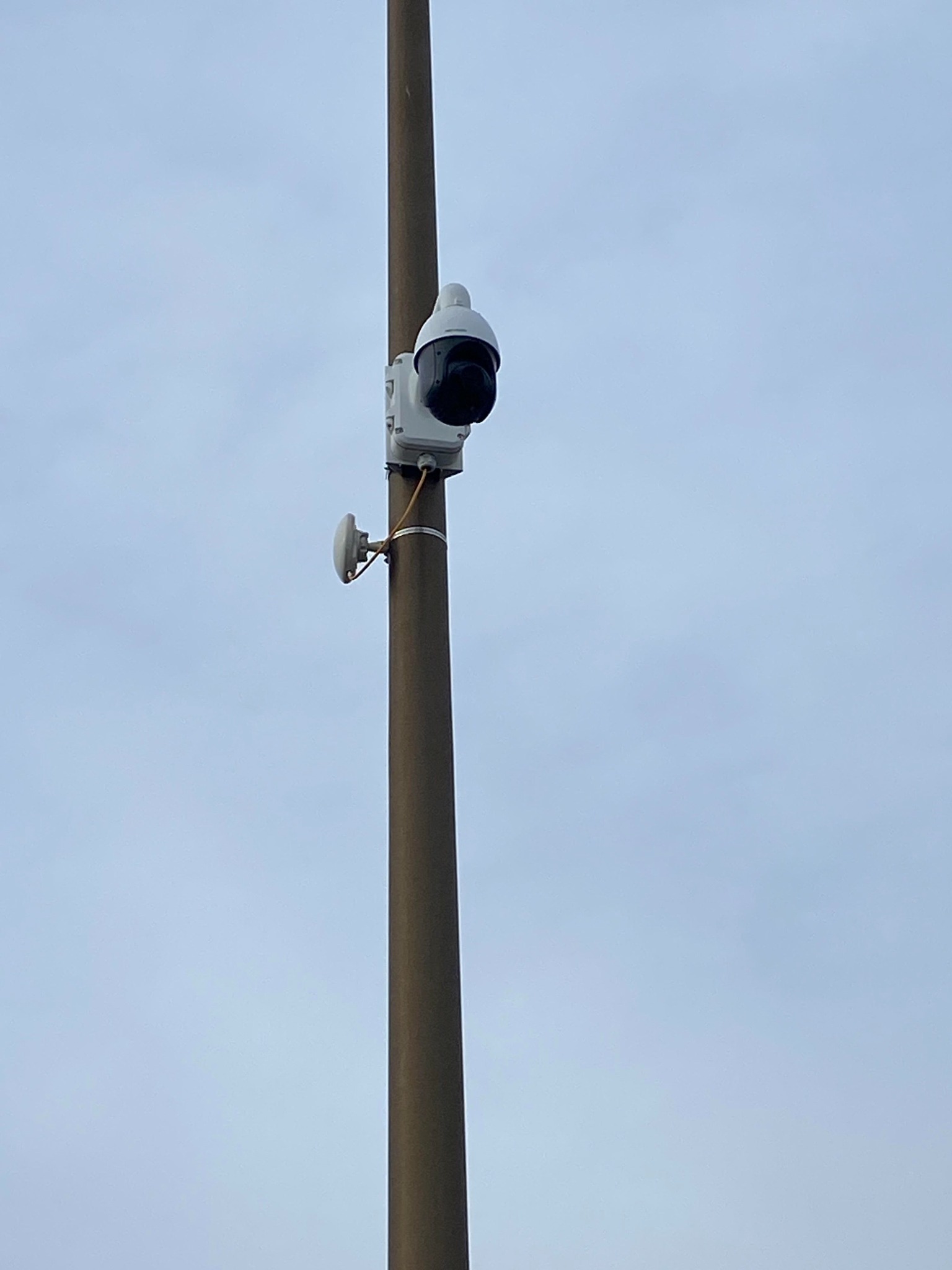 Kamera kopułkowa CCTV zamontowana na brązowym słupie z anteną Wi-Fi przymocowaną poniżej, na tle jasnego, pochmurnego nieba.
