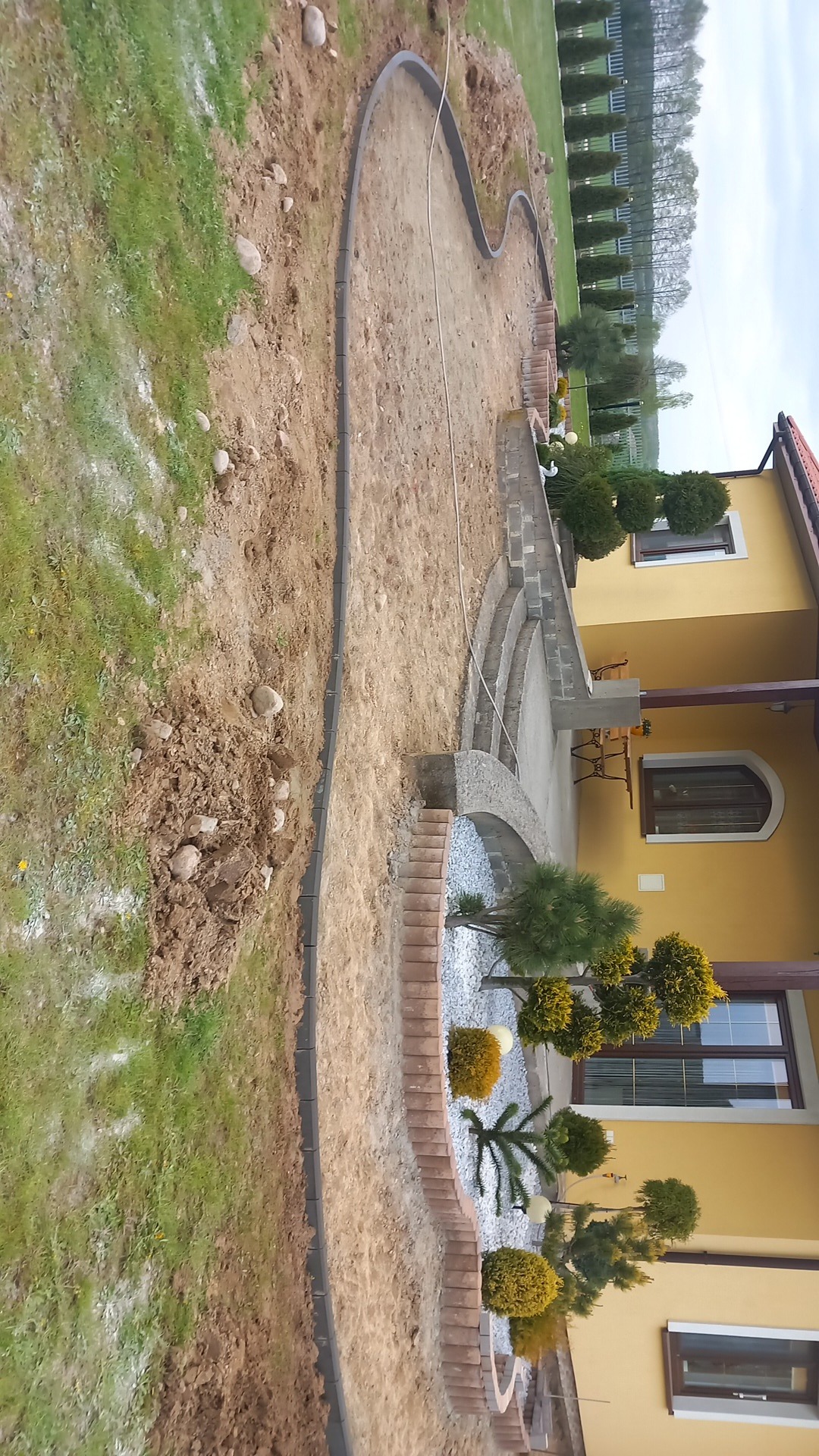 Krawężnik z kostki brukowej oddzielający trawnik od terenu przygotowanego pod ścieżkę, z widocznymi elementami architektury ogrodowej i fragmentem elewacji budynku.