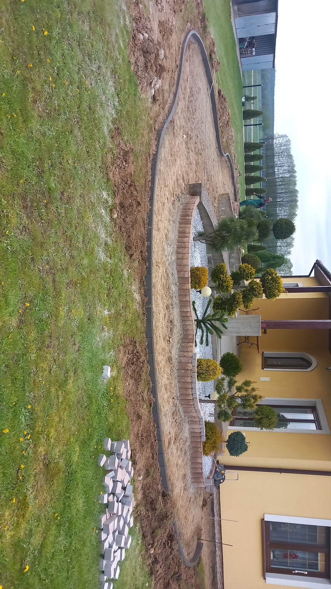 Kształtowanie terenu pod ogród z obrzeżami z kostki i przygotowaną podbudową, widoczne kostki do ułożenia na trawniku, w tle żółty budynek.