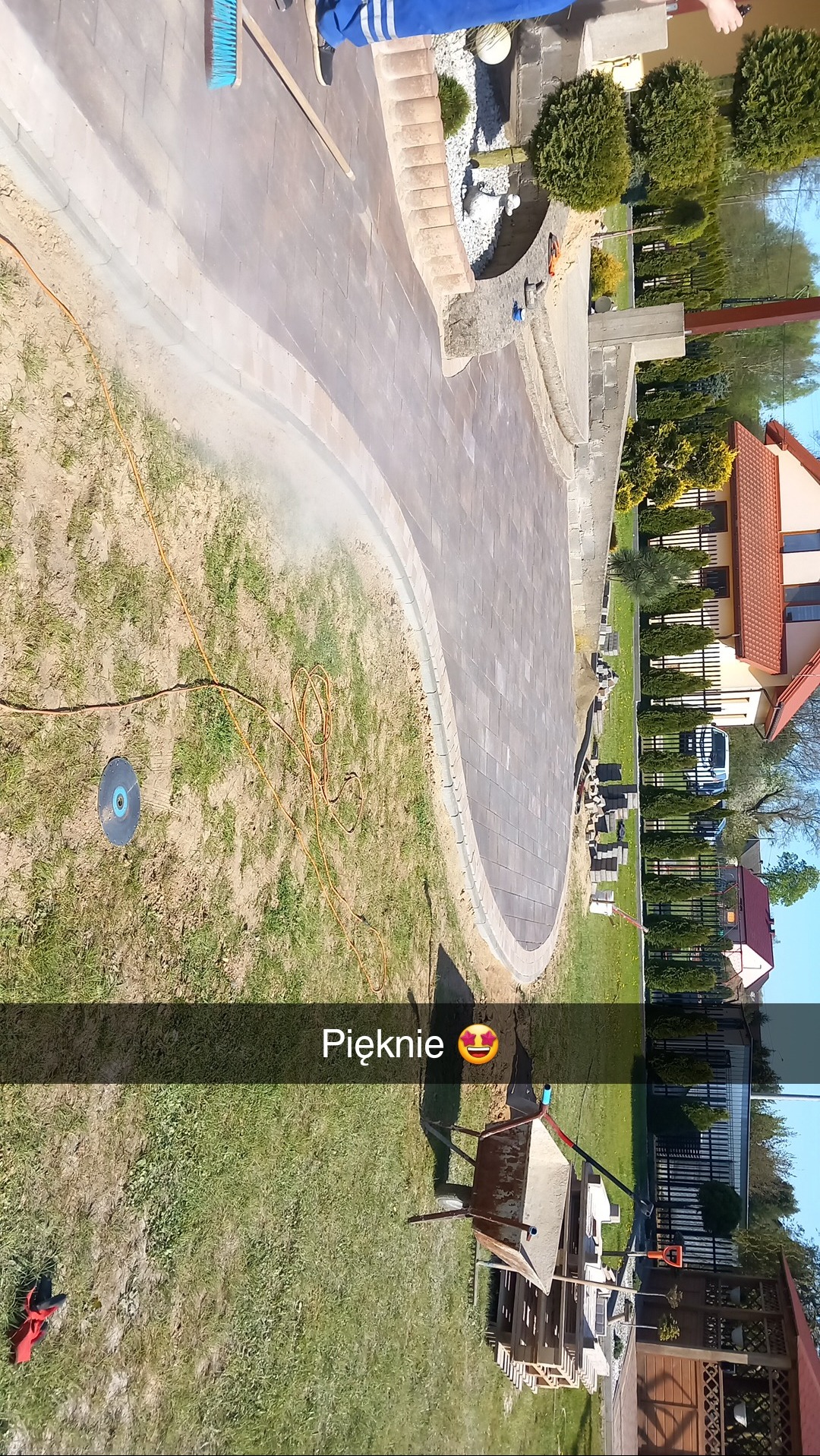 Krzywoliniowy chodnik z kostki, częściowo ułożony, z widocznymi narzędziami i materiałami budowlanymi, w tle dom i ogród.