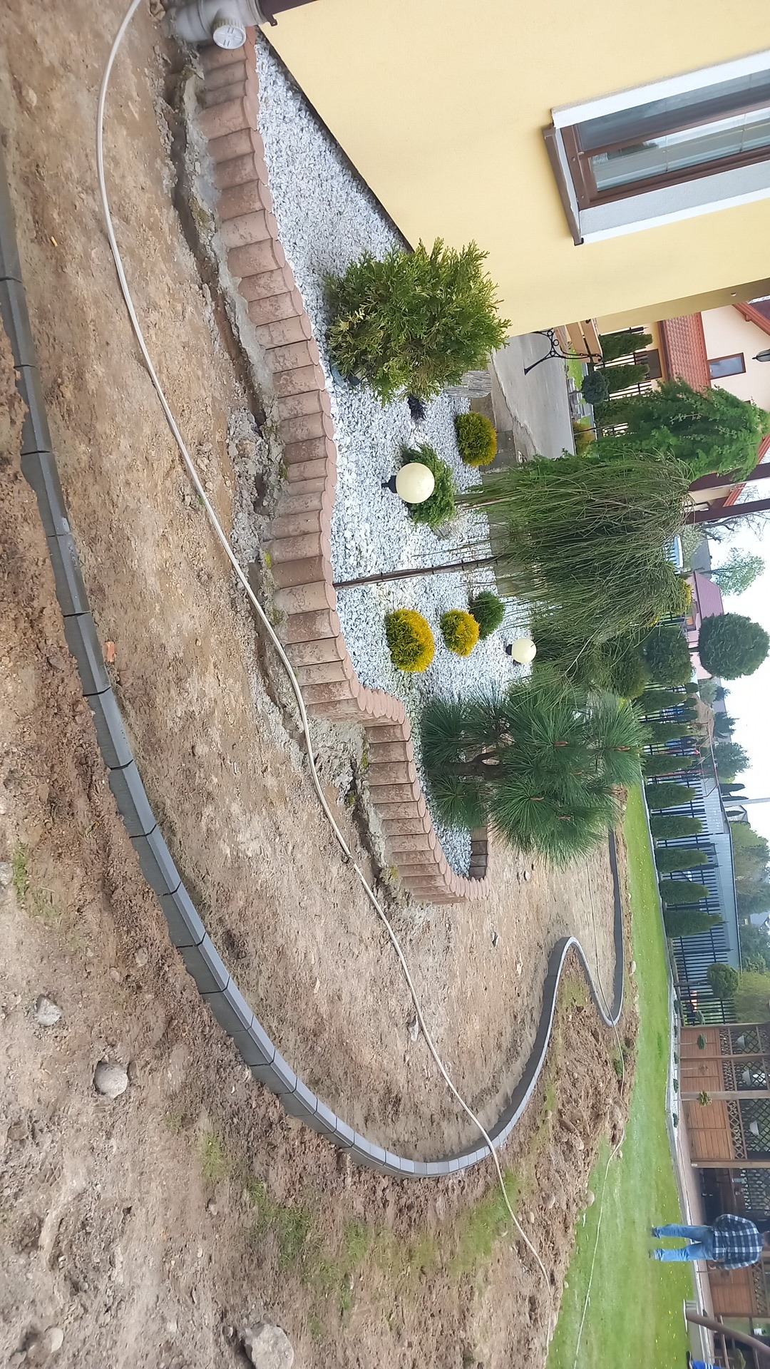 Krawężniki z kostki betonowej wyznaczające ścieżkę w ogrodzie, obok rabata z kamykami, krzewami i lampami ogrodowymi, widoczne elementy domu i ogrodzenia.