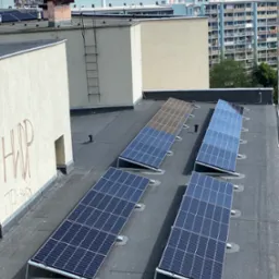 Panele słoneczne zamontowane na płaskim dachu budynku, widok z góry na tle miejskiego krajobrazu z blokami.