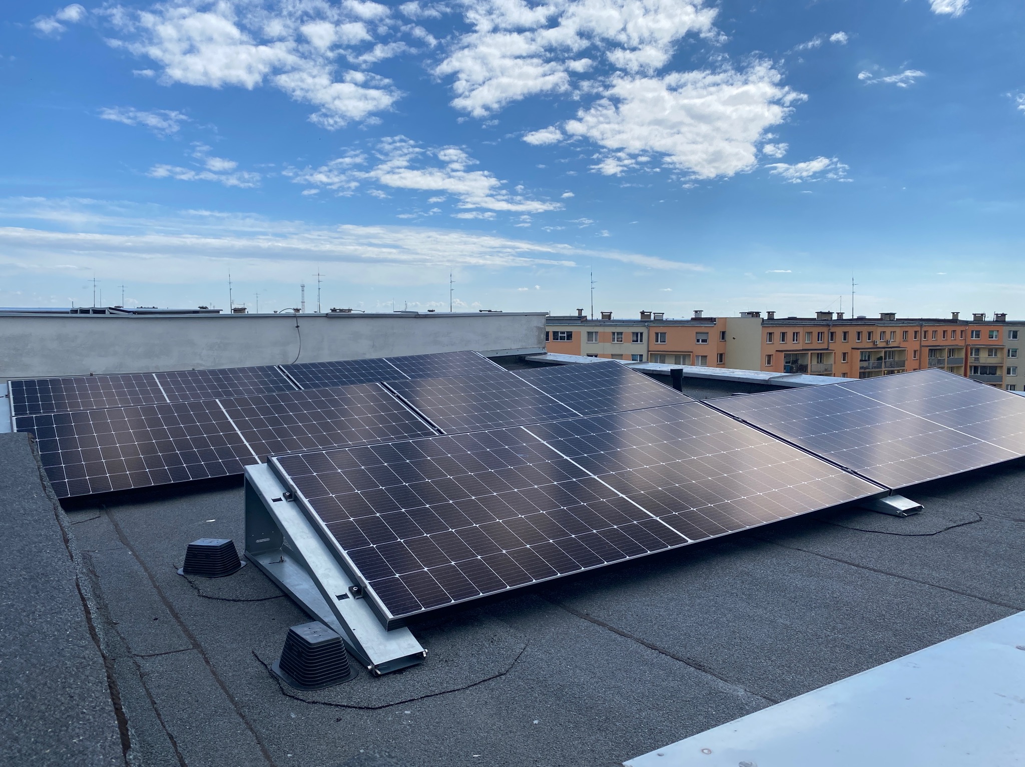 Panele słoneczne zamontowane na płaskim dachu budynku wielorodzinnego, widoczne elementy mocujące i przewody, błękitne niebo z chmurami w tle.