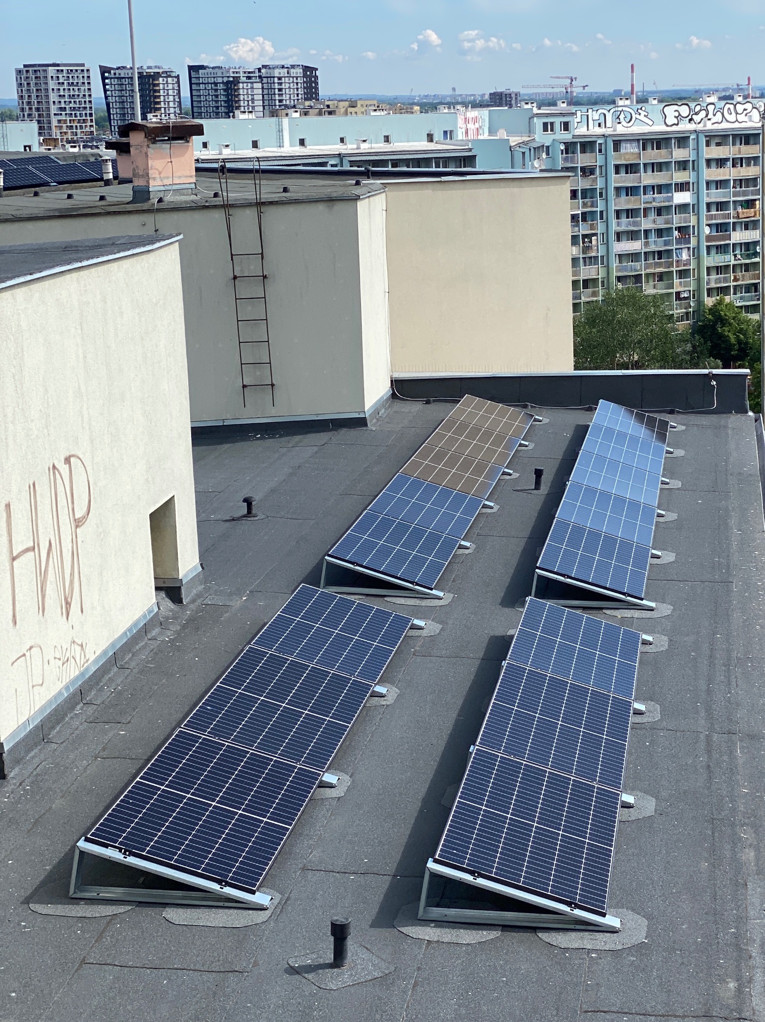 Panele słoneczne zamontowane na płaskim dachu budynku, widok z góry na tle miejskiego krajobrazu z blokami.