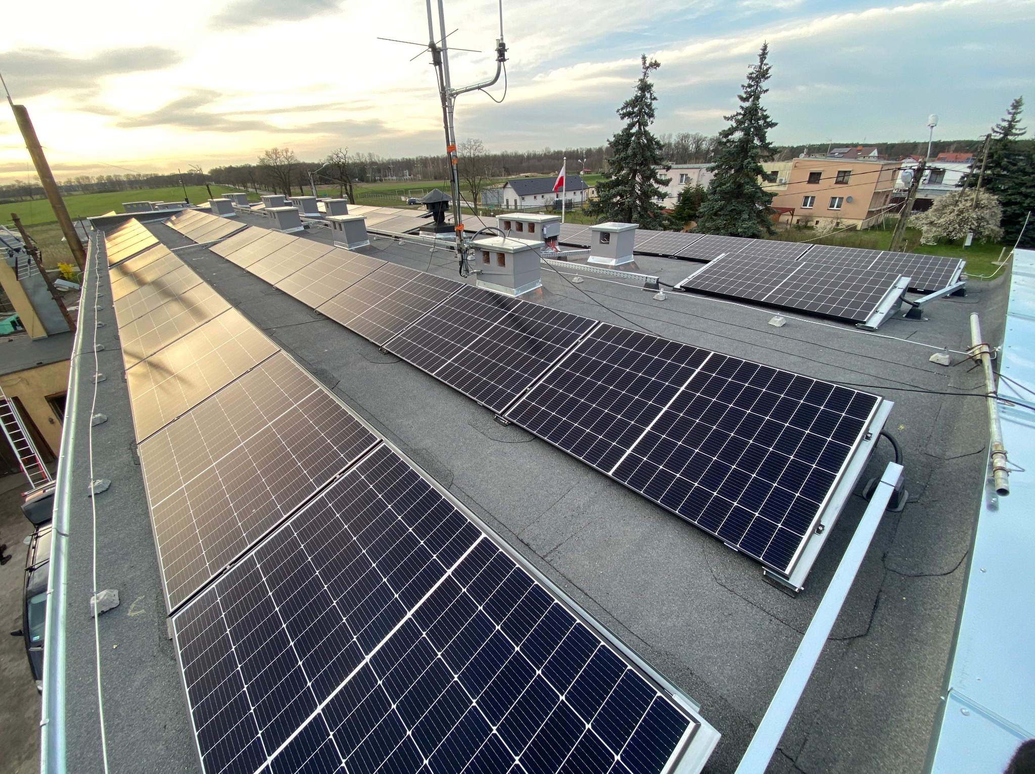 Panele słoneczne na płaskim dachu budynku z widoczną anteną, kominami wentylacyjnymi i okoliczną zabudową w tle.