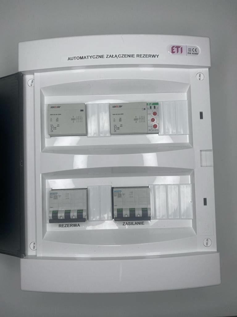 SZR: Biała skrzynka z automatycznym załączaniem rezerwy, widoczne moduły sterujące i wyłączniki zasilania oraz rezerwy.