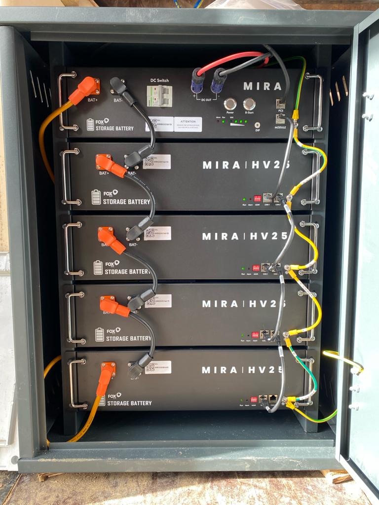 Rząd akumulatorów energii MIRA HV25 w szafie, podłączonych kablami z pomarańczowymi końcówkami i zielono-żółtymi przewodami uziemiającymi.