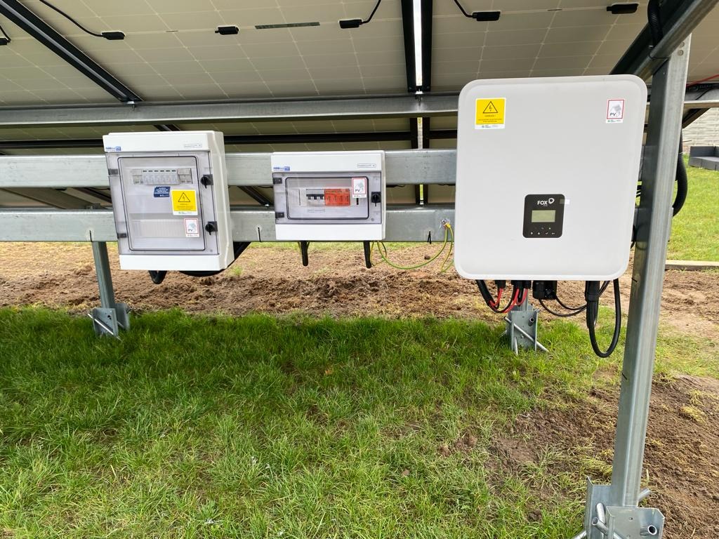 Instalacja paneli słonecznych: widok na białą skrzynkę inwertera FoxESS i dwie skrzynki zabezpieczeń na stalowej konstrukcji, na tle trawnika.