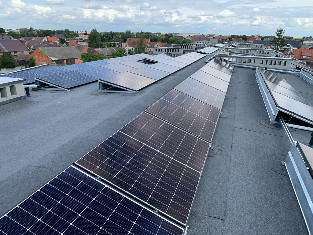 Rozległa instalacja fotowoltaiczna na płaskim dachu budynku, widok z góry, w tle zabudowa miejska i pochmurne niebo.