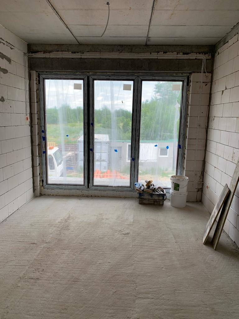 Surowe wnętrze w trakcie wykończenia: widoczne ściany z bloczków, okno z widokiem na zewnątrz, narzędzia w skrzynce, kable elektryczne pod sufitem.