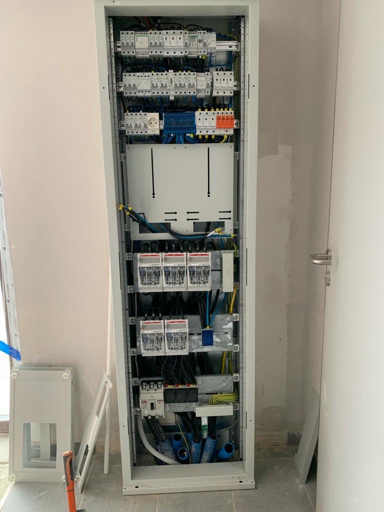 Otwarta szafa elektryczna z widocznymi bezpiecznikami, przewodami i elementami instalacji, dodatkowa pusta obudowa szafy obok.