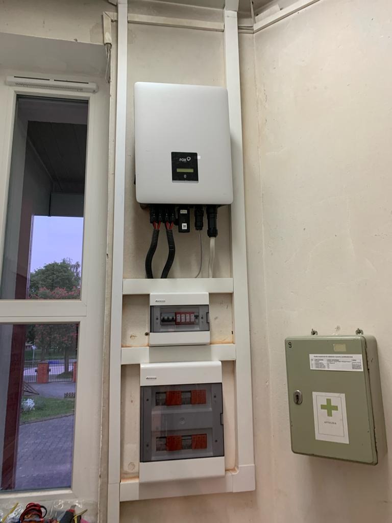 Biały falownik zamontowany na ścianie obok okna i skrzynek elektrycznych, z widoczną apteczką pierwszej pomocy po prawej stronie.