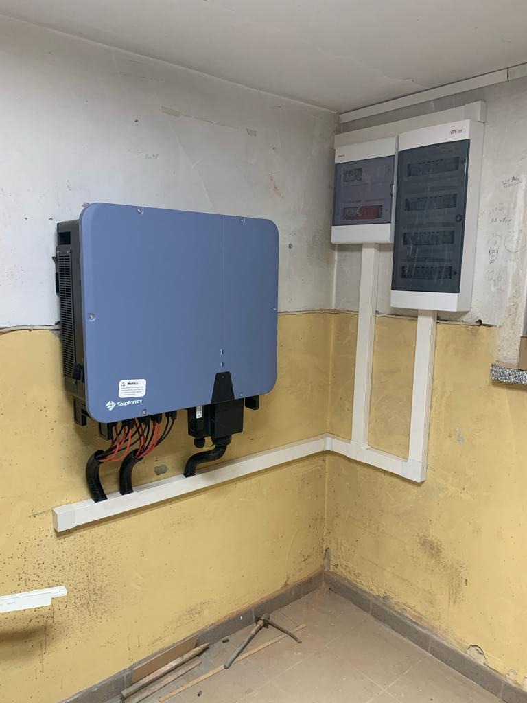 Szary falownik fotowoltaiczny marki Solplanet zamontowany na ścianie w pomieszczeniu z żółtą, niedokończoną ścianą, widoczne kable i rurki instalacyjne.