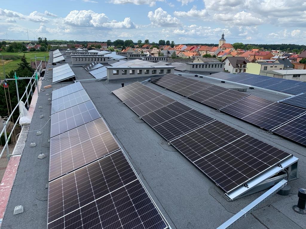 Rząd paneli słonecznych na płaskim dachu budynku z widokiem na miasto z kościołem w tle, częściowo zamontowane, widoczne zabezpieczenia na krawędzi dachu.