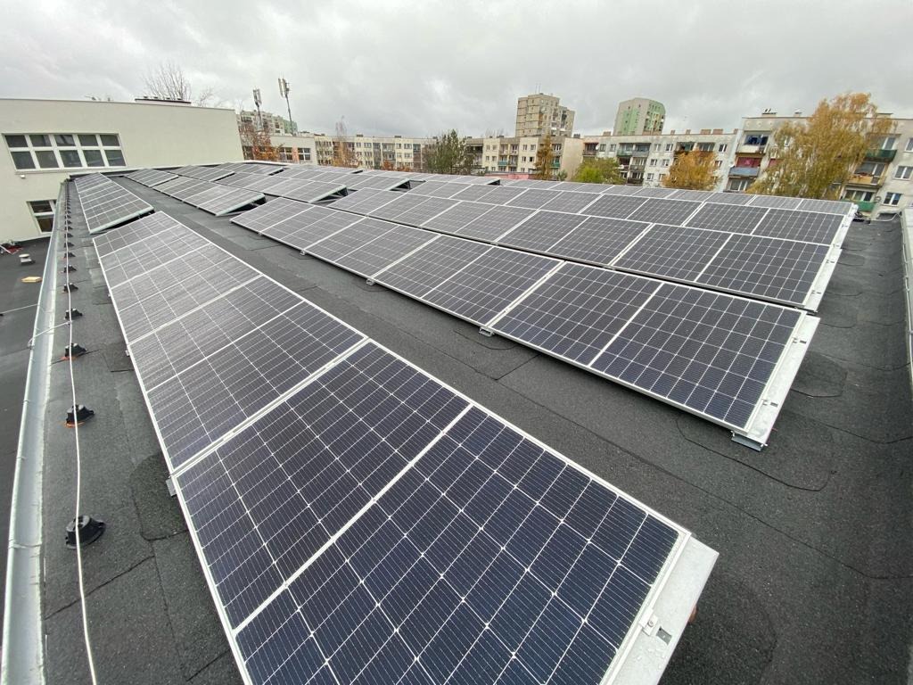 Instalacja paneli słonecznych na płaskim dachu budynku, widok z góry, szare niebo w tle.