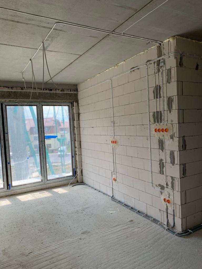 Surowy stan wnętrza z widoczną instalacją elektryczną: kable w peszlach prowadzone po ścianach z betonu komórkowego i pod sufitem, puszki elektryczne zamontowane w ścianie.