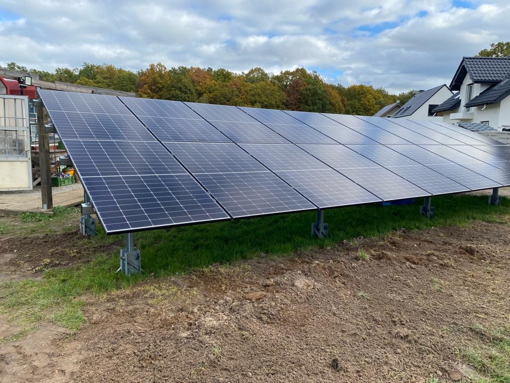 Zestaw paneli słonecznych na metalowych wspornikach, zainstalowany na trawiastym terenie z widocznym budynkiem mieszkalnym w tle.