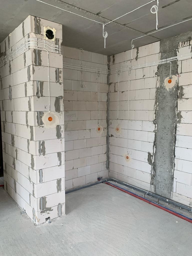 Surowy stan pomieszczenia z instalacją elektryczną w peszlach na ścianach z betonu komórkowego i instalacją wodną w podłodze, widoczne otwory pod gniazdka.