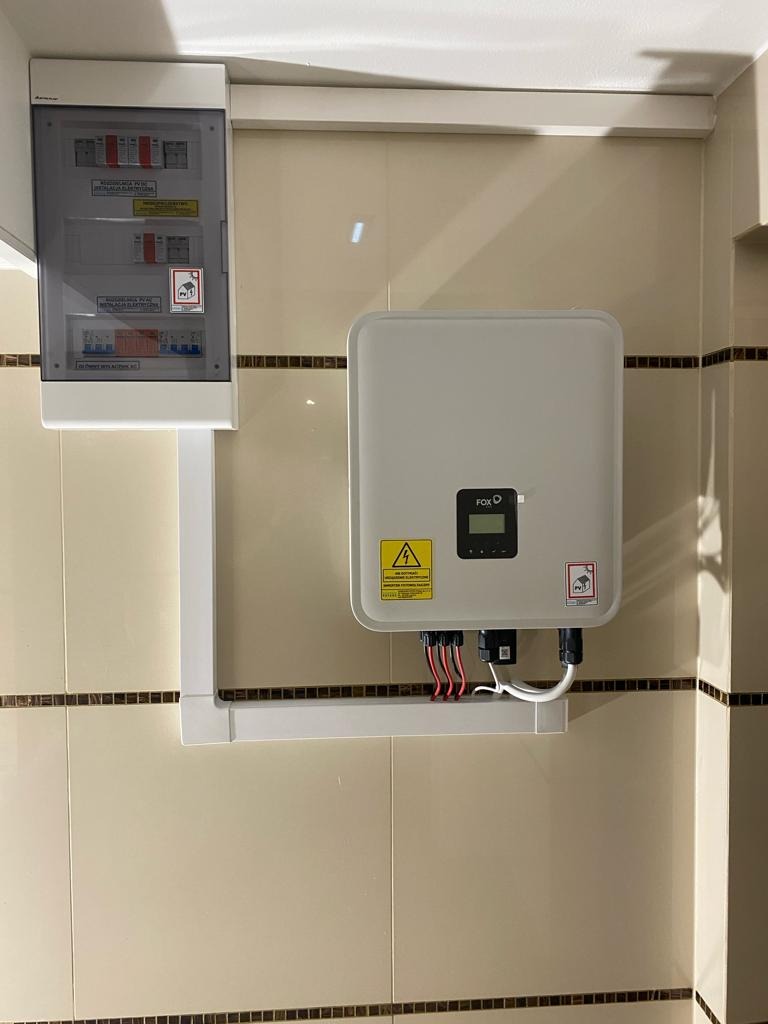 Zamontowany inwerter marki FOX z podłączonymi kablami, obok skrzynka bezpiecznikowa, na tle beżowych płytek z ciemnym paskiem.