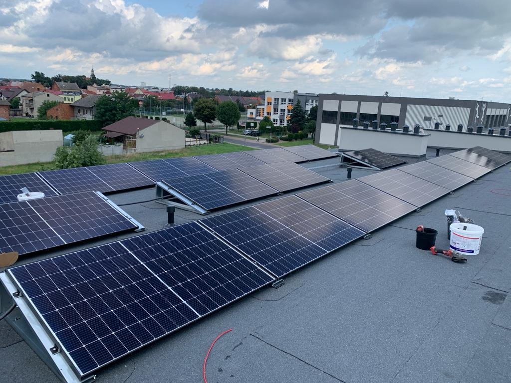 Panele słoneczne na dachu budynku, widok z góry, w tle zabudowa miejska i pochmurne niebo. Widoczne narzędzia i wiadro z klejem. Część paneli jeszcze nie zamontowana.