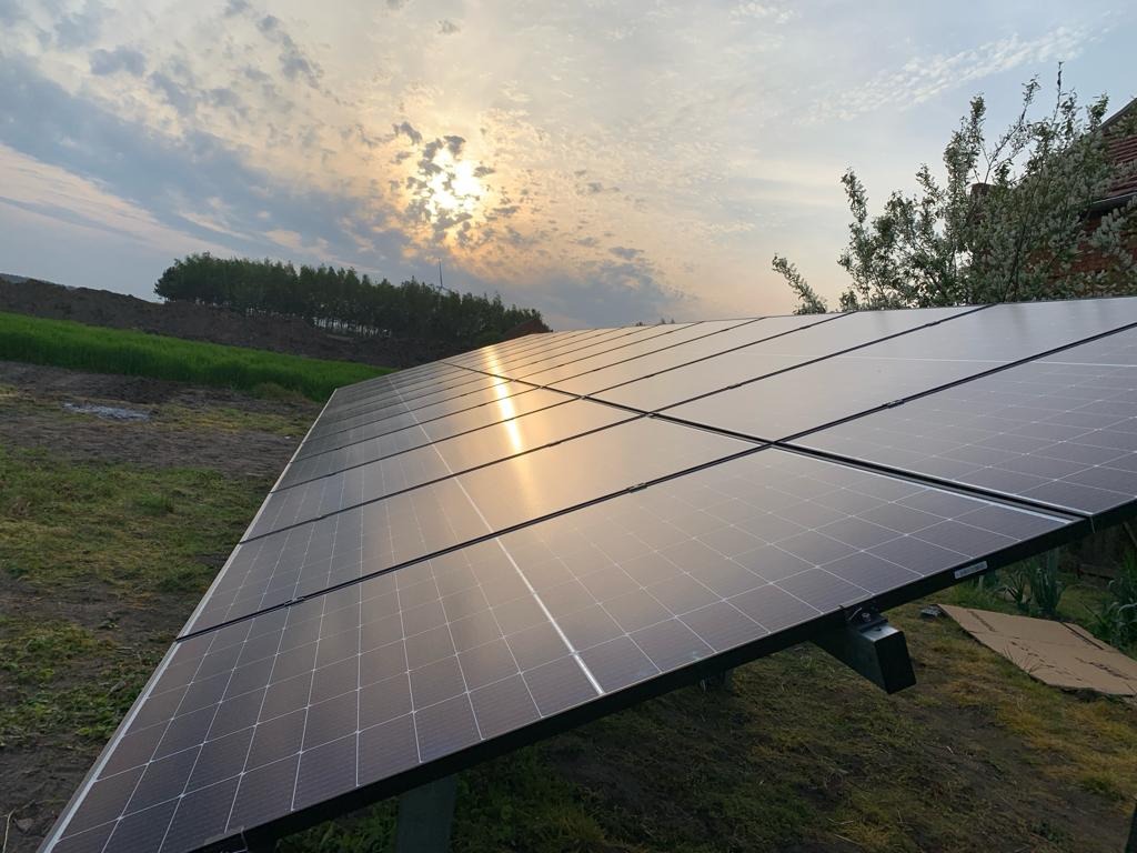 Instalacja fotowoltaiczna na trawiastym terenie, odbicie zachodzącego słońca w panelach, widok na horyzont z drzewami i częściowo zachmurzone niebo.