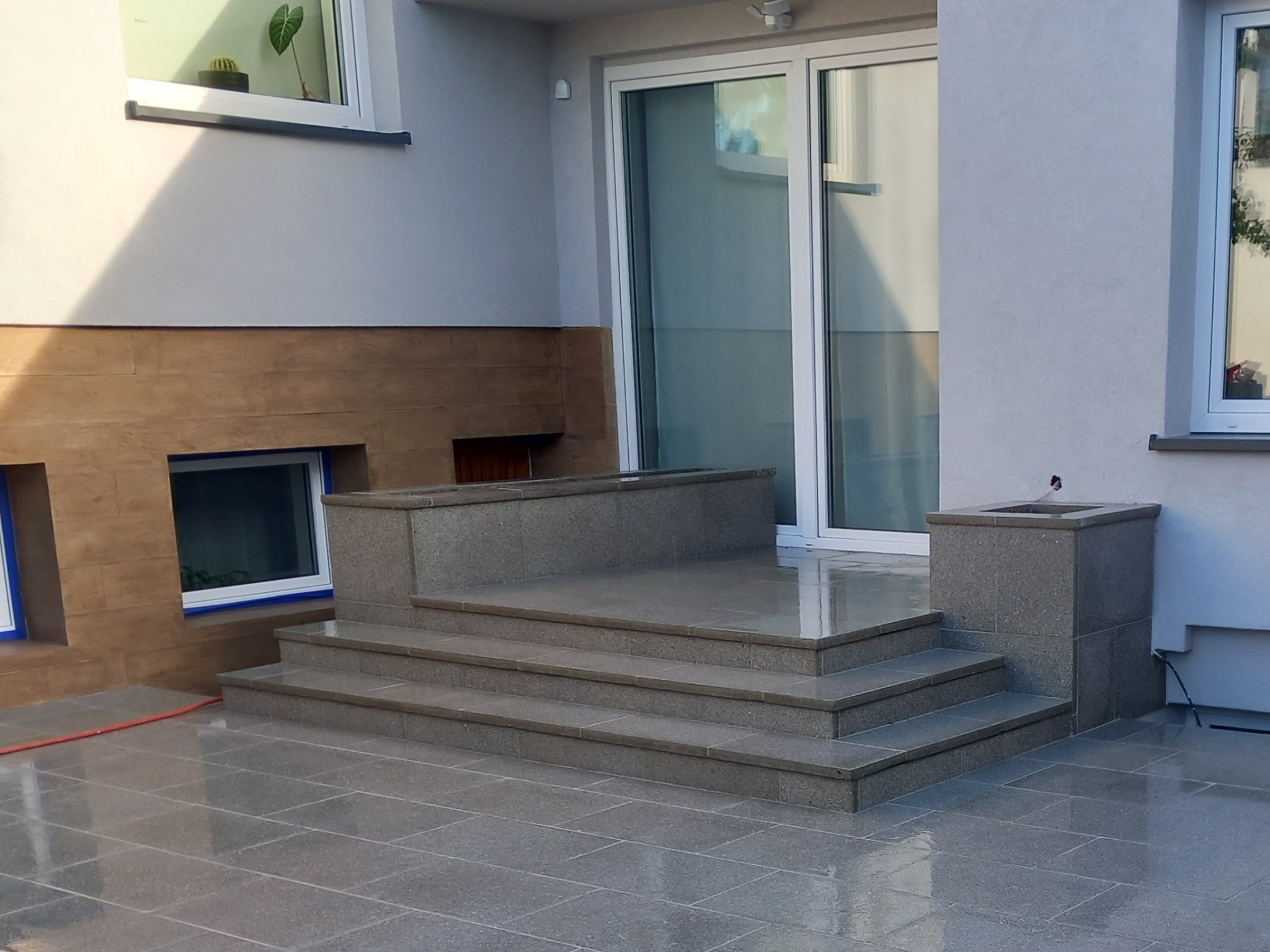 Granitowe schody wejściowe z podestem i donicą, prowadzące do przeszklonych drzwi tarasowych, otoczone jasnoszarą elewacją z elementami dekoracyjnymi w kolorze brązu, na pierwszym planie...