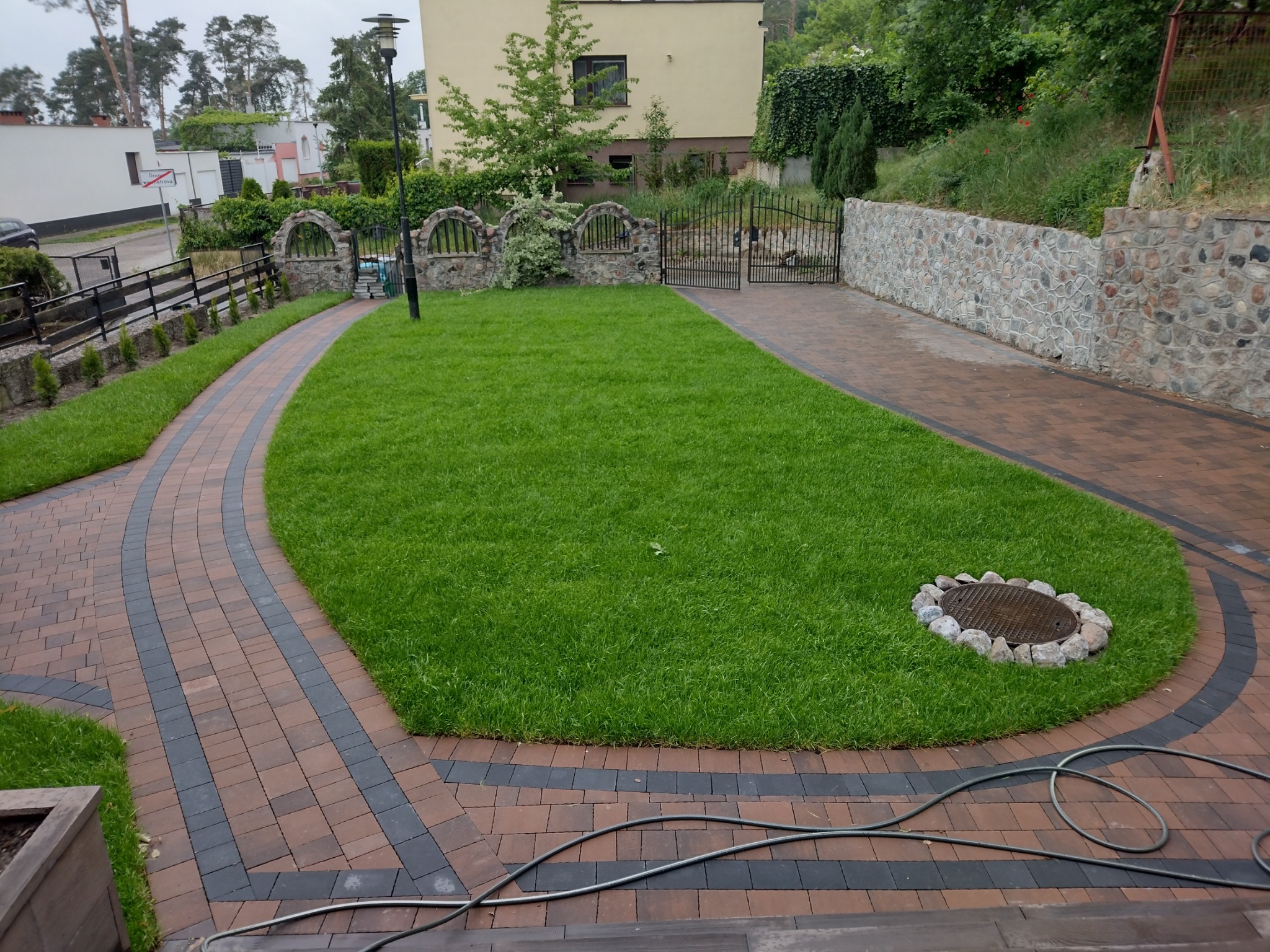 Brązowo-czarna kostka brukowa tworząca ścieżkę i podjazd wokół trawnika z okrągłą studzienką kanalizacyjną otoczoną kamieniami, w tle dom i kamienny murek.