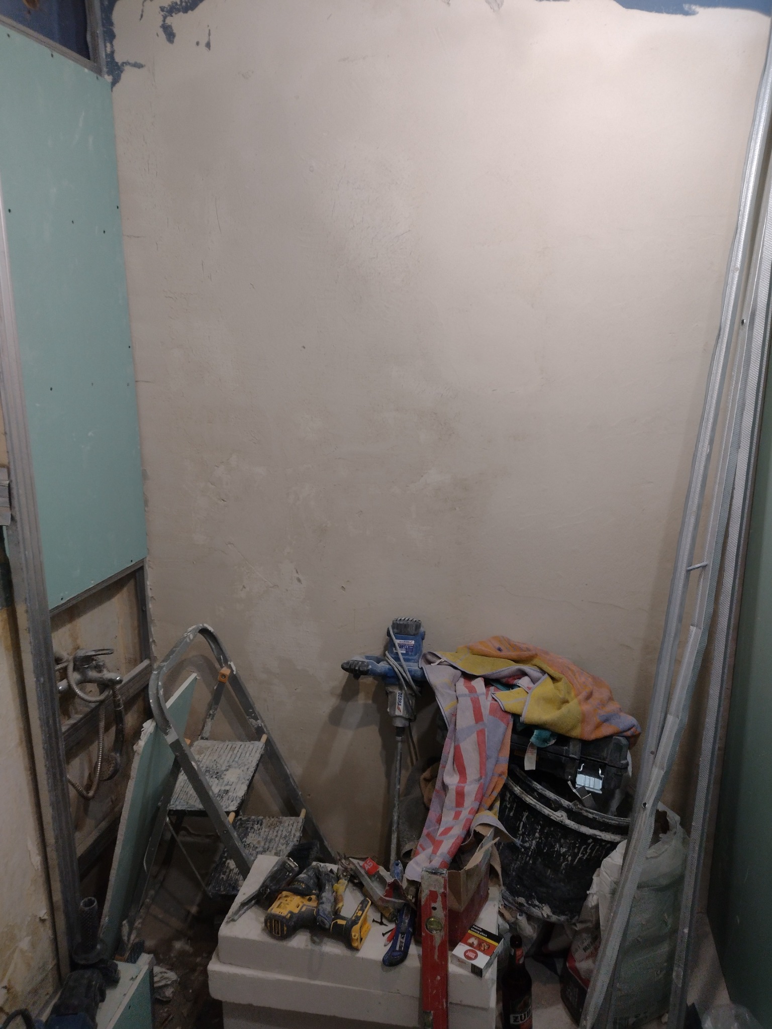 Niedokończona ściana po nałożeniu gładzi, widoczny stelaż pod płytę karton gips, drabina, mieszadło do gipsu, narzędzia oraz akcesoria budowlane.
