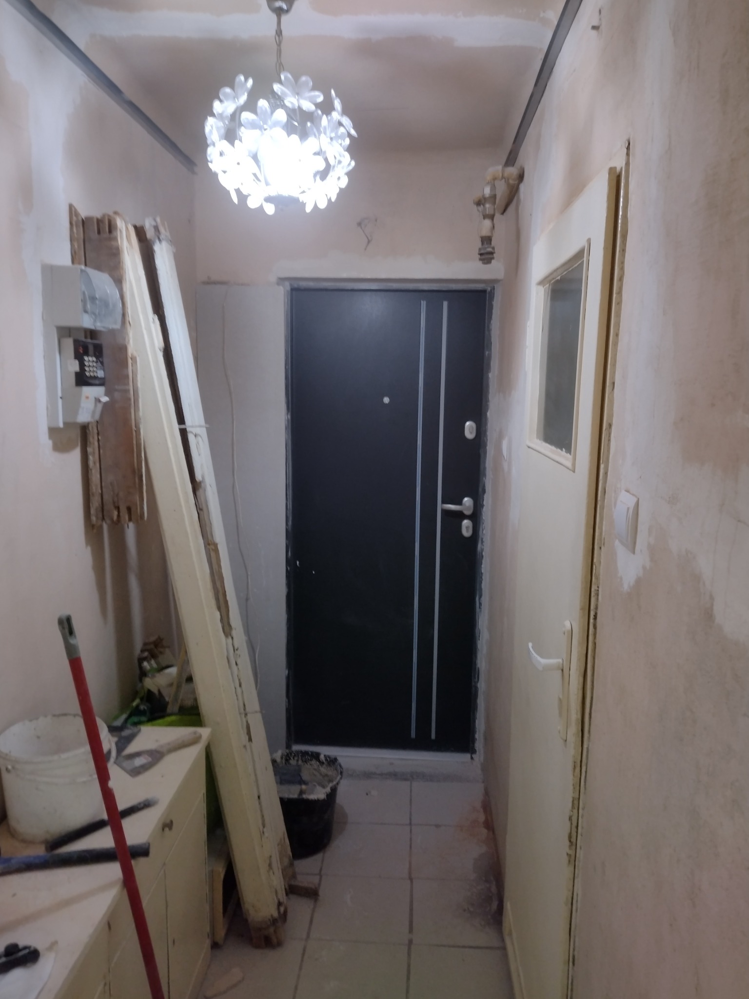 Korytarz w trakcie remontu: ściany przygotowane pod malowanie, widoczne przewody, stara ościeżnica oparta o ścianę, wisząca lampa w kształcie kwiatów.