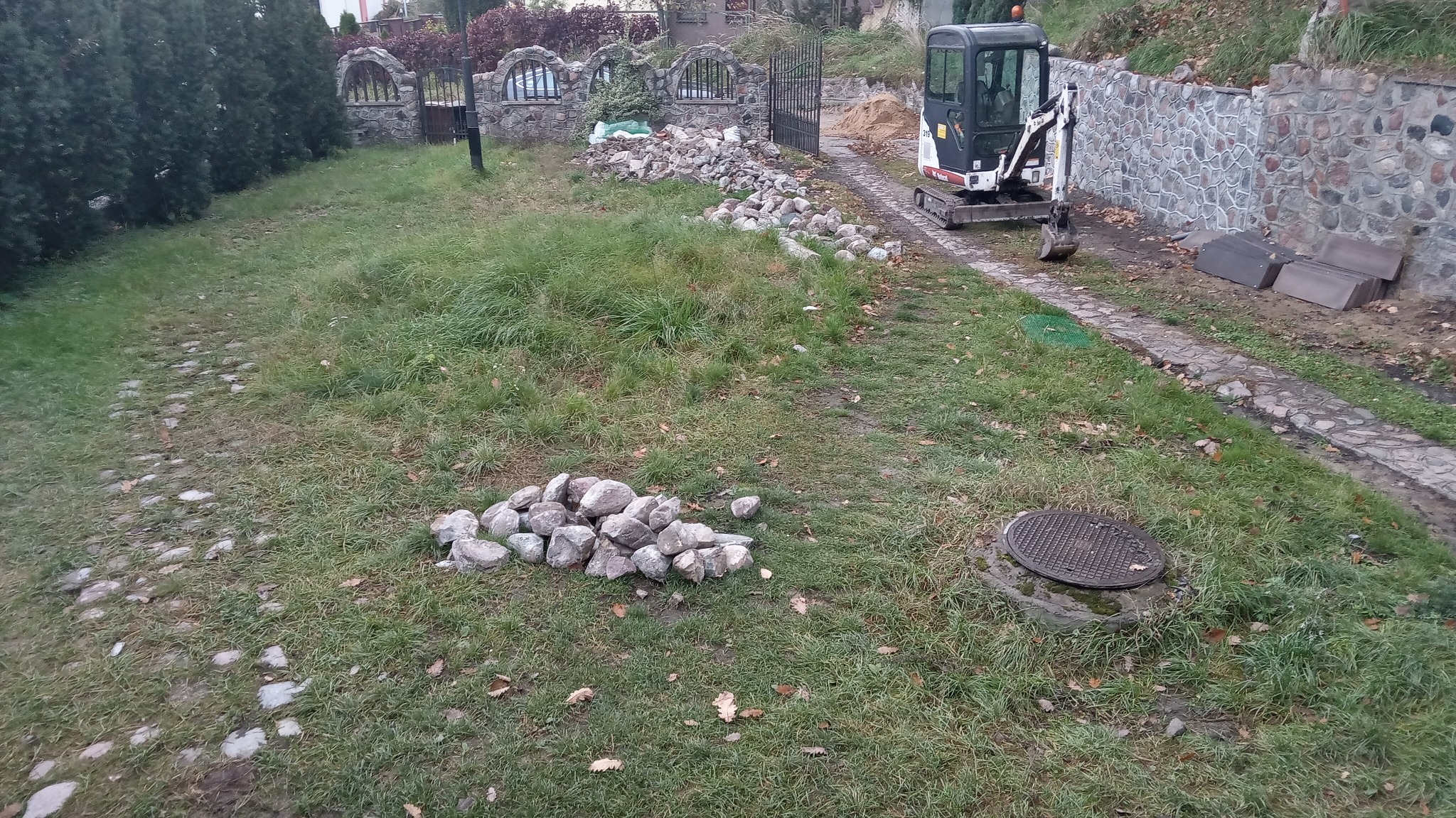 Teren przed posesją w trakcie przygotowania do prac brukarskich; widoczny mini-koparka, stos kamieni i fragment ułożonej ścieżki z kamienia naturalnego.