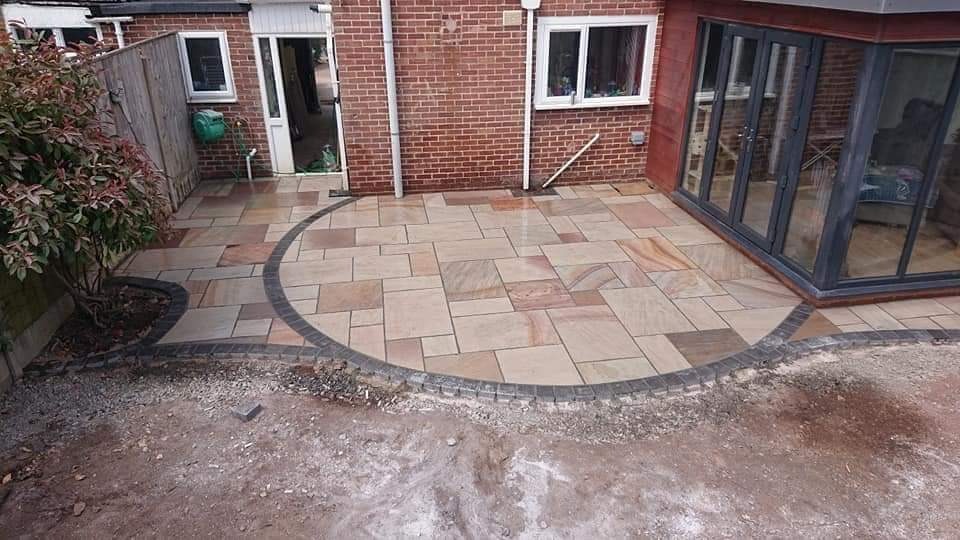 Nowo ułożony taras z piaskowca z obramowaniem z ciemnej kostki, przylegający do ceglanego budynku z przeszklonymi drzwiami, widoczny fragment rabaty z krzewem.