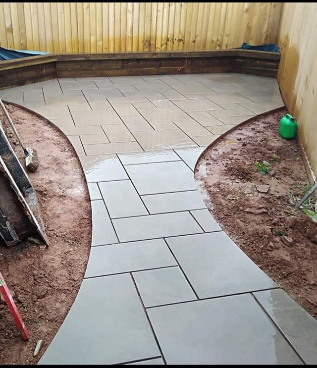 Ułożona ścieżka z szarych płyt na zewnątrz, mokra od wody, z widocznymi narzędziami i zielonym kanistrem w tle drewniany płot.