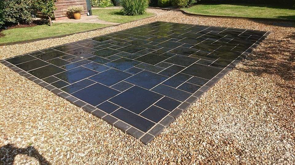 Ułożona, ciemna kostka na podwórku otoczona drobnym żwirem, z widocznym fragmentem trawnika i niewielkiego budynku w tle.