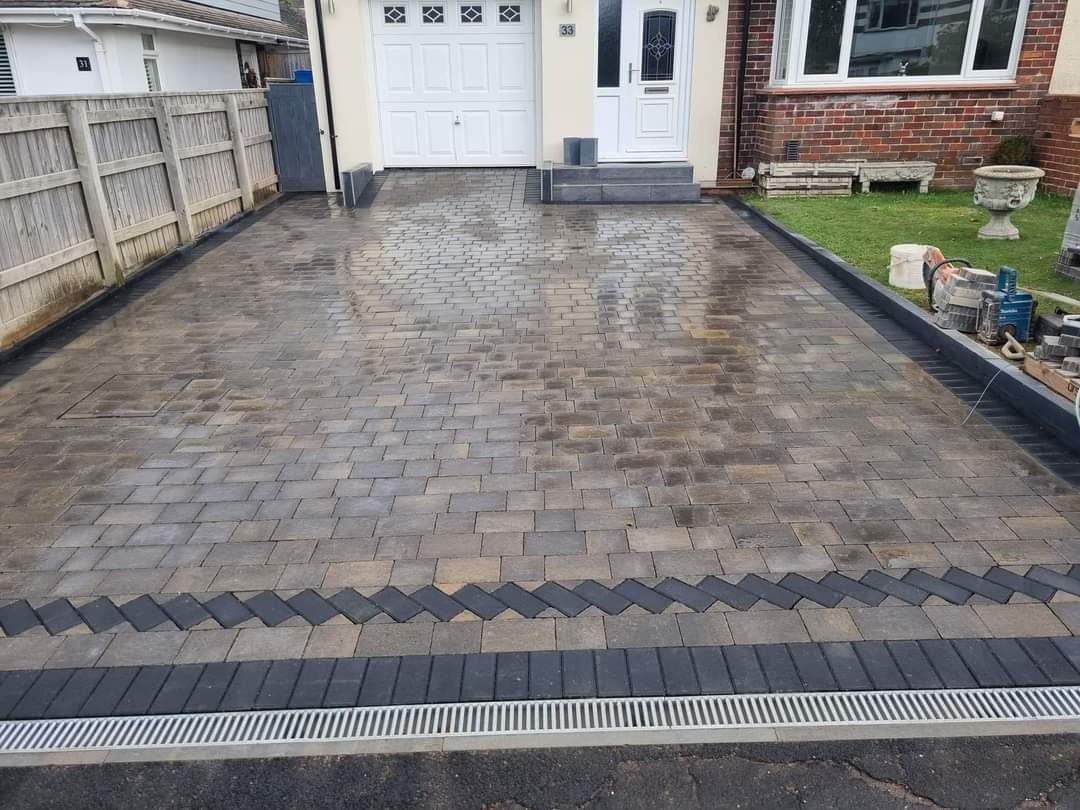 Ułożona kostka brukowa w odcieniach szarości i brązu, z dekoracyjnym pasem w kształcie zygzaka i odpływem liniowym, prowadząca do garażu i domu z ceglaną fasadą.