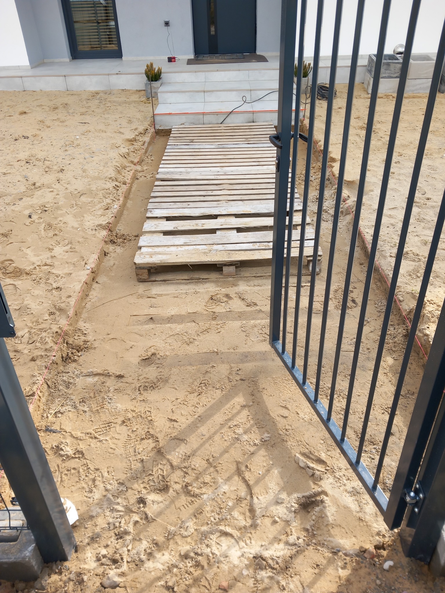 Tymczasowa ścieżka z palet prowadząca do wejścia domu, widok przez otwartą, metalową bramę w trakcie prac ziemnych i przygotowania podłoża, widoczne wytyczone krawędzie.