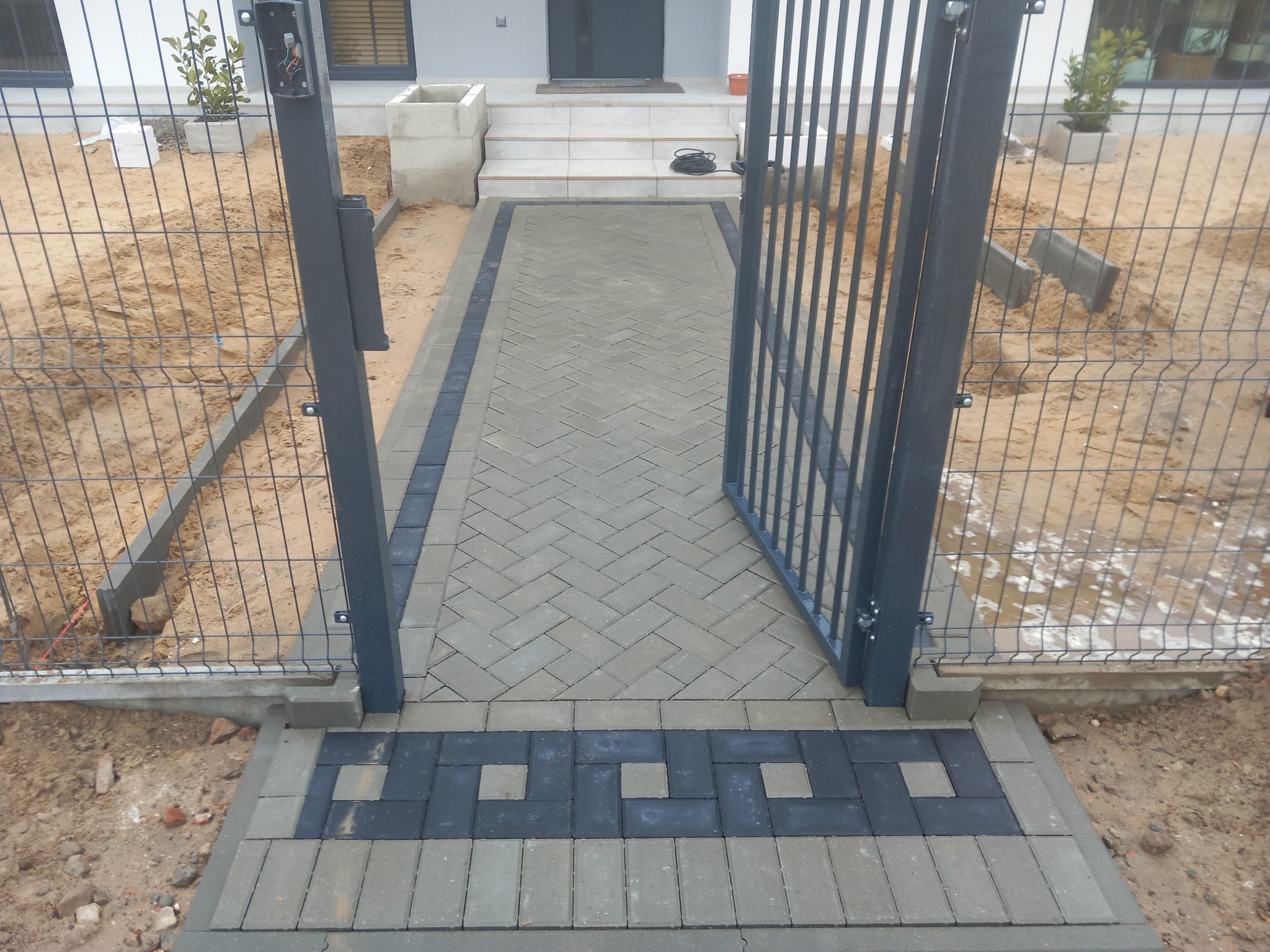 Ścieżka z jasnoszarej kostki brukowej ułożonej w jodełkę, obramowana ciemniejszą kostką, prowadząca od furtki z metalowej siatki do wejścia domu z białymi schodami.