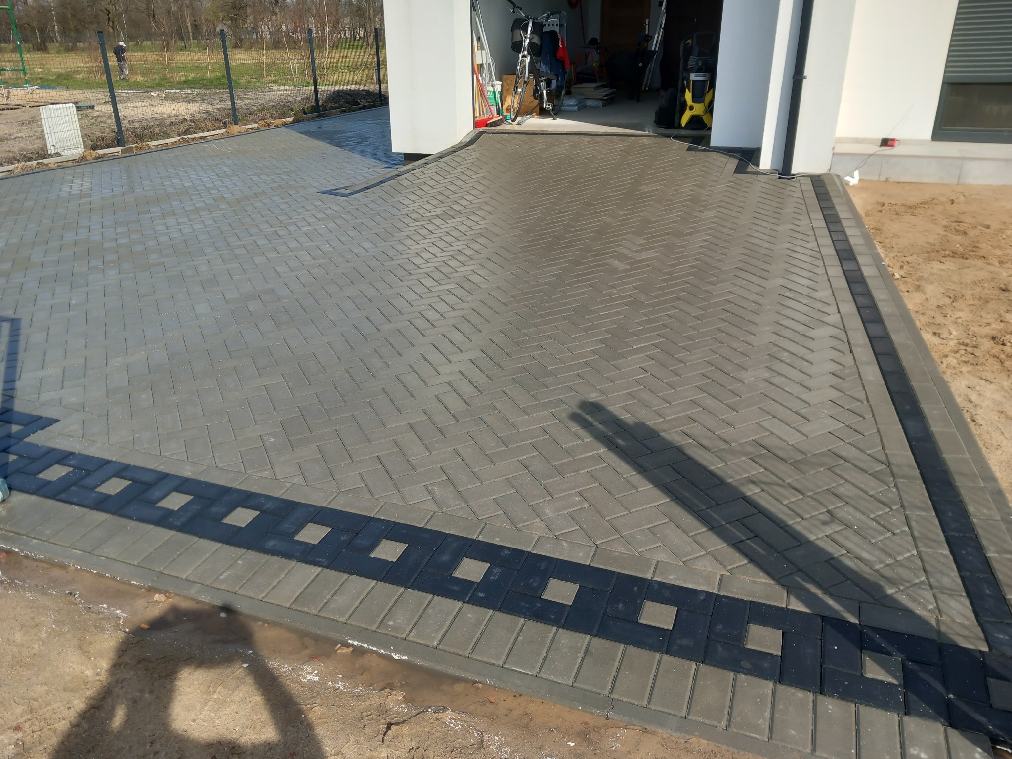 Ułożona kostka brukowa w jodełkę na podjeździe, z ciemniejszym obramowaniem z kwadratowymi wstawkami, widoczny cień osoby fotografującej.