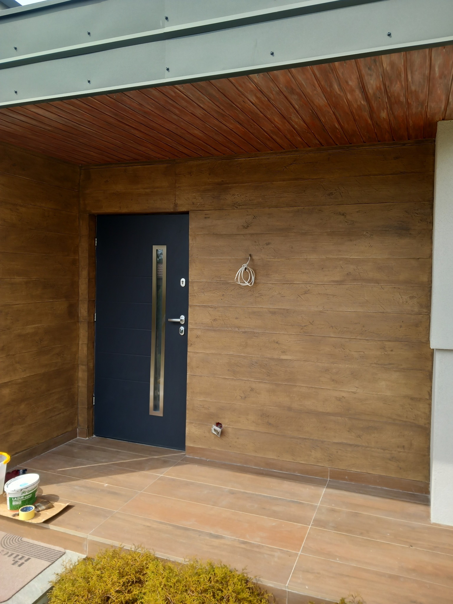 Elewacja domu z efektem drewnianych desek, nowoczesne drzwi wejściowe z przeszkleniem, brązowa terakota na podłodze, widoczne puszki z lazurą do drewna i przewody elektryczne wystające ze ściany.