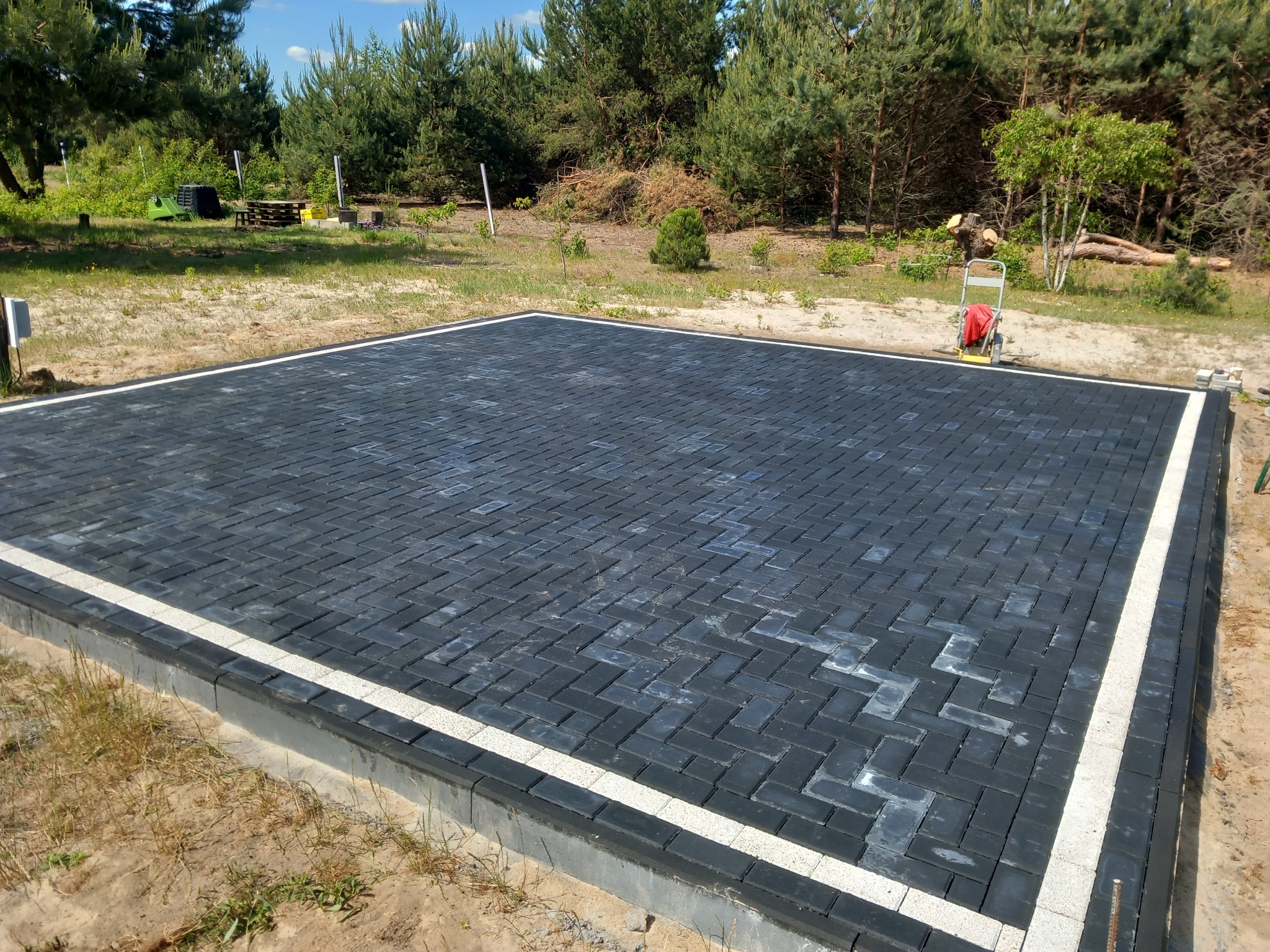 Nowo ułożona, czarna kostka brukowa w kształcie prostokątów, obramowana białym pasem, otoczona piaszczystym terenem i drzewami w tle.