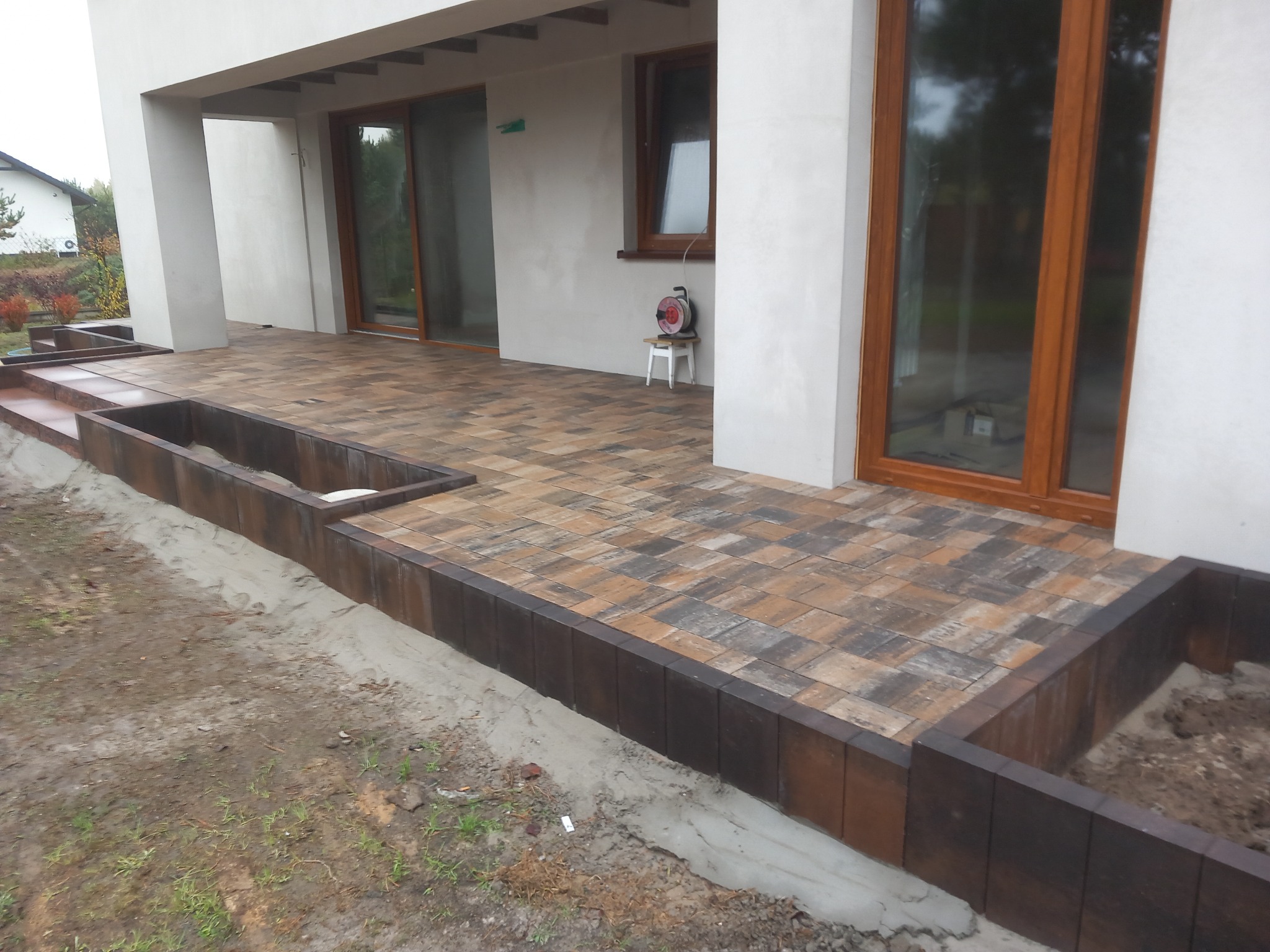 Nowoczesny taras z kostki w odcieniach brązu i szarości, obrzeża z ciemnego drewna, widoczne donice bez roślin, biała elewacja budynku z dużymi oknami z brązowymi ramami, na tarasie stoi mały...