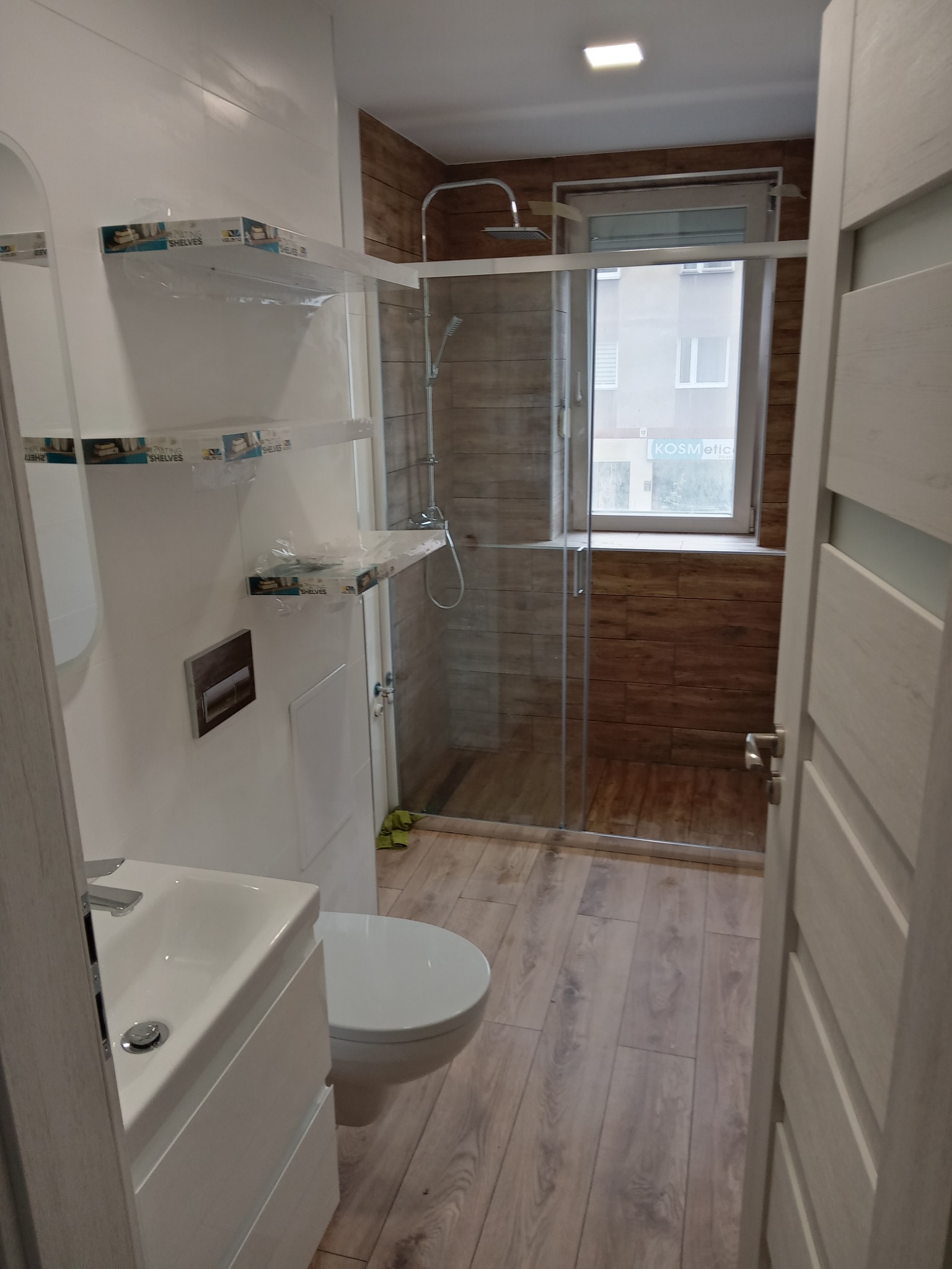 Nowoczesna łazienka z kabiną prysznicową typu walk-in, umywalką z szafką w kolorze białym, podwieszaną toaletą i drewnopodobną podłogą, z widocznymi półkami w folii ochronnej.